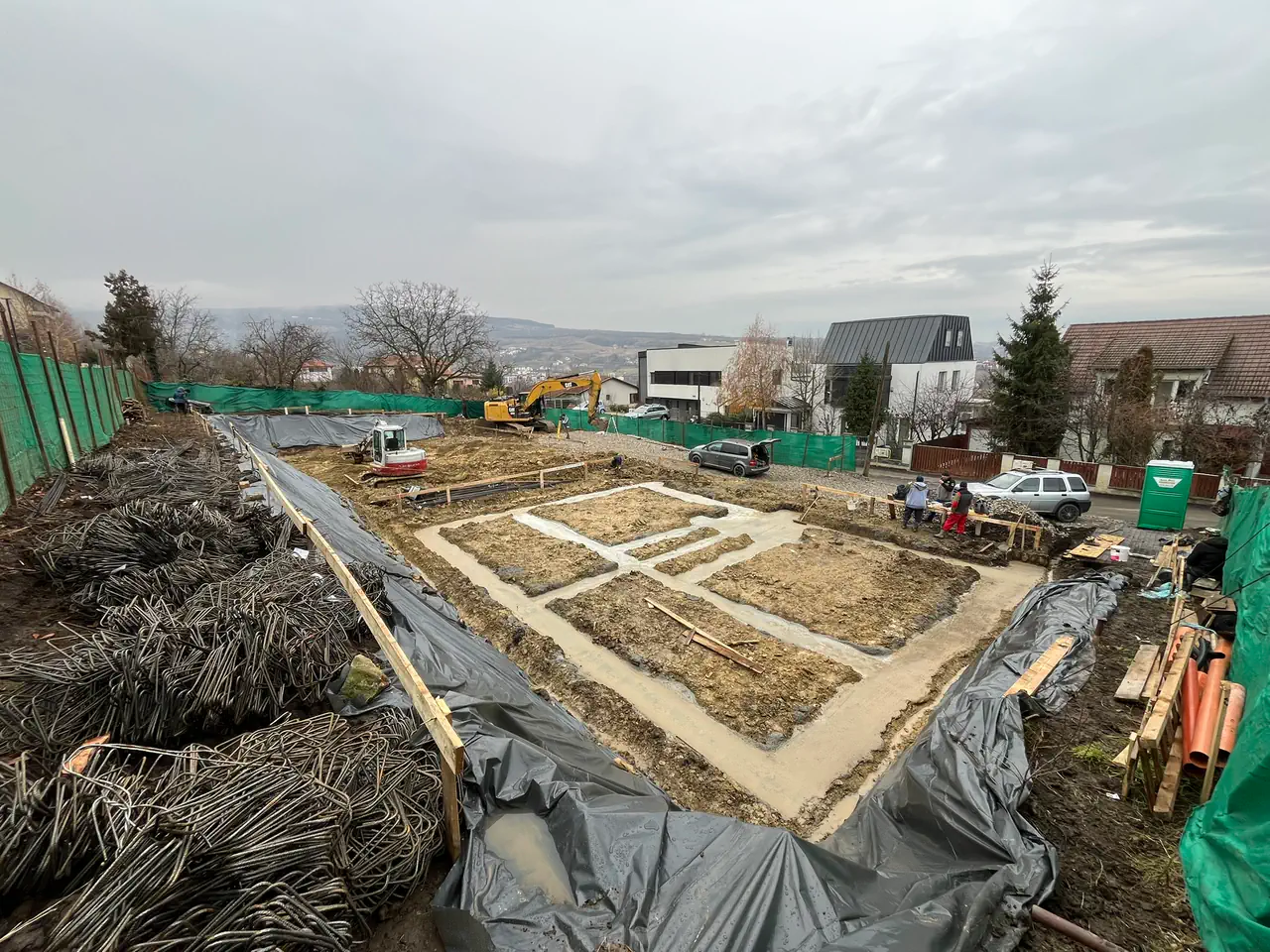 Vedere de ansamblu asupra săpăturii, cu betonul turnat în tranșeele fundațiilor și colacii de armătură alături.
