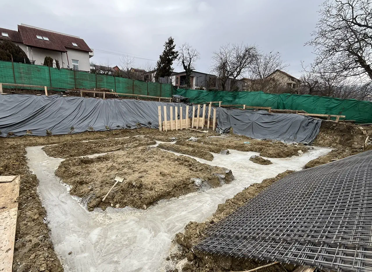 Tranșeele fundațiilor cu betonul de egalizare turnat și plasele de armătură pregătite pe margine.
